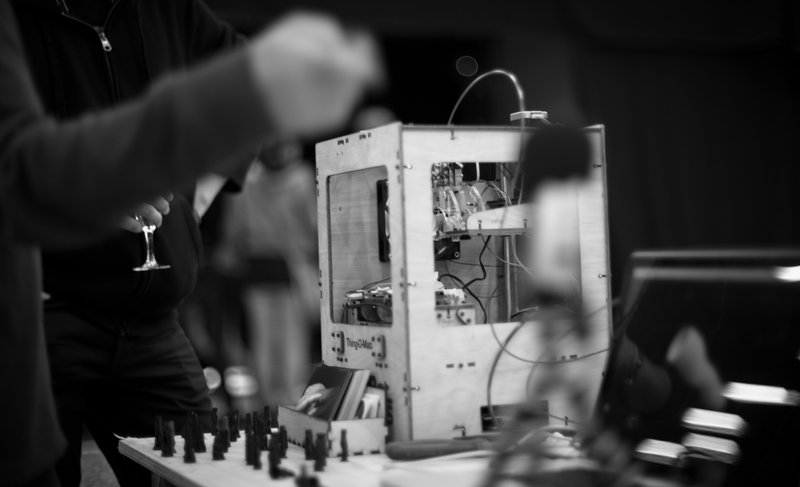 A Thing-o-Matic 3D printer on a table behind a microphone, with vague outlines of people standing behind and in front of the table an arm pointing downwards; underneath the arm is a partially visible hand holding a glass of wine.